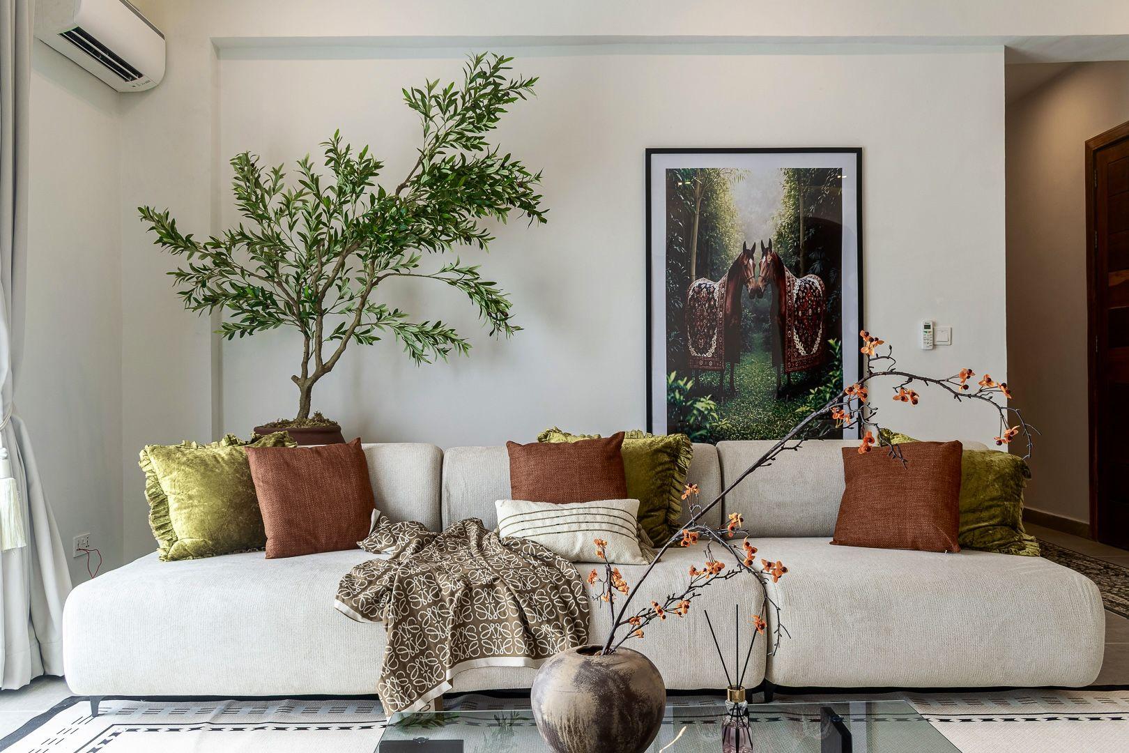 Living room with a cream sofa, olive and rust cushions, an olive tree and framed artwork of two dressed horses