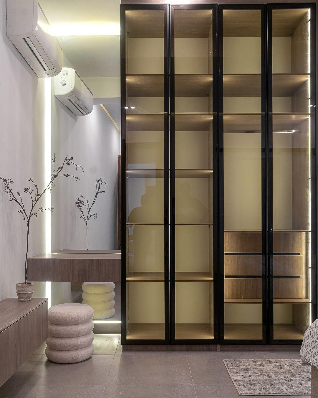 Glass fronted wardrobe wall with lit shelving beside a boucle stool